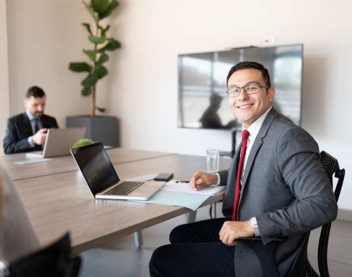 Smiling man in glasses and a suit sitting at a boardroom table with a colleague working in the background